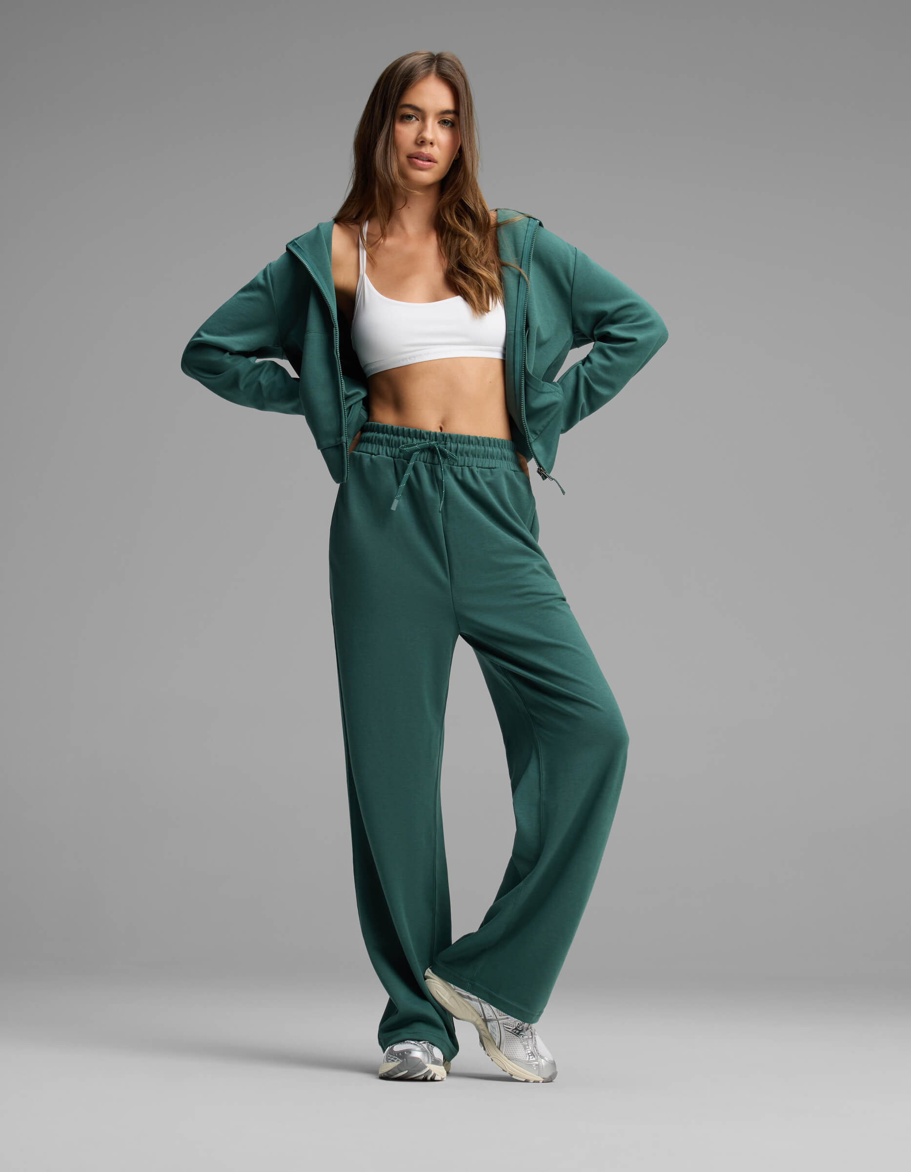 A woman poses in the Cozy Earth Women's Versa Zip-Down Hoodie & Straight Leg Pant Set, featuring a green hue, paired with a white sports bra and white sneakers, standing against a plain gray background.|Color:Uniform