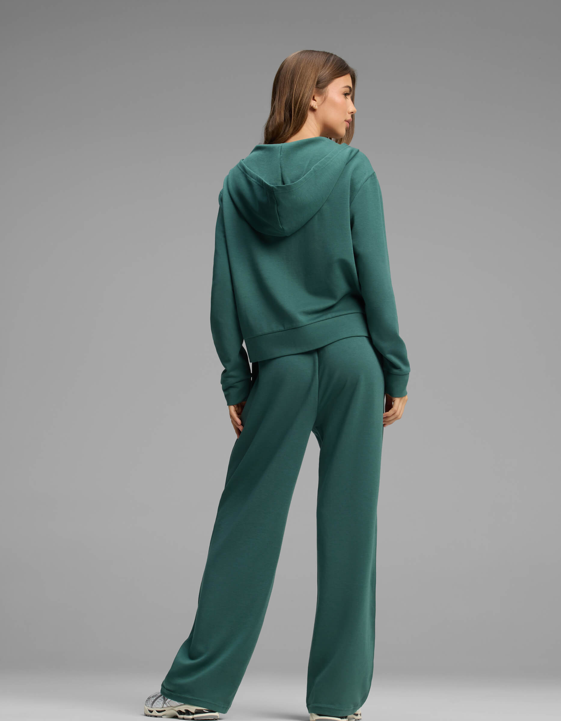 A person stands facing away, wearing the Cozy Earth Women's Versa Zip-Down Hoodie & Straight Leg Pant Set in green with white sneakers against a plain gray background.|Color:Uniform