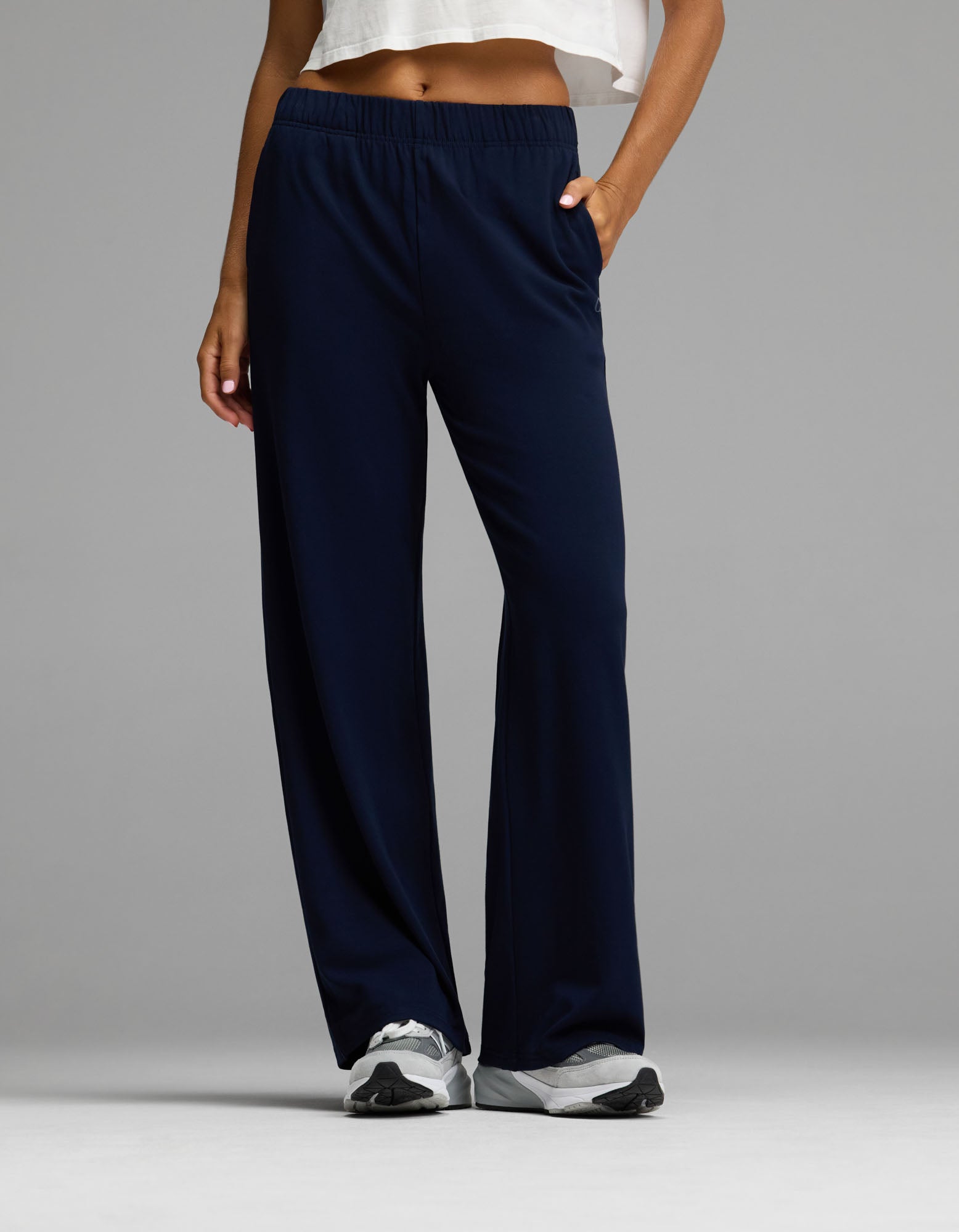 A person wearing the Cozy Earth Women’s Brushed Bamboo Wide Leg Pull-On Pant in Navy, paired with a white cropped top and white sneakers, stands with one hand in their pocket against a plain gray background.