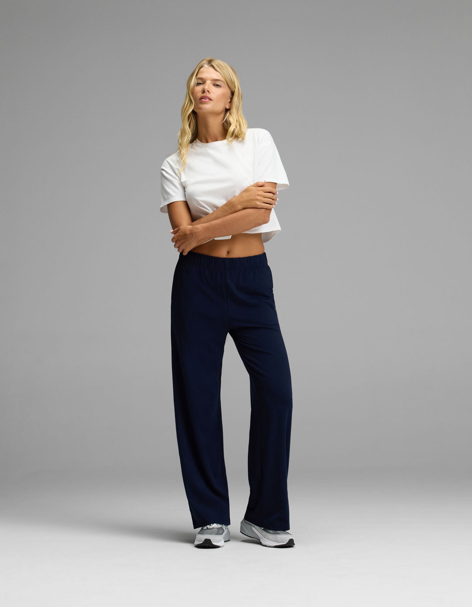 A person stands against a plain background, wearing a white cropped t-shirt, white sneakers, and the Cozy Earth Women’s Brushed Bamboo Wide Leg Pull-On Pant in dark blue. Their long blonde hair falls over their shoulders as they cross their arms and look off to the side.
