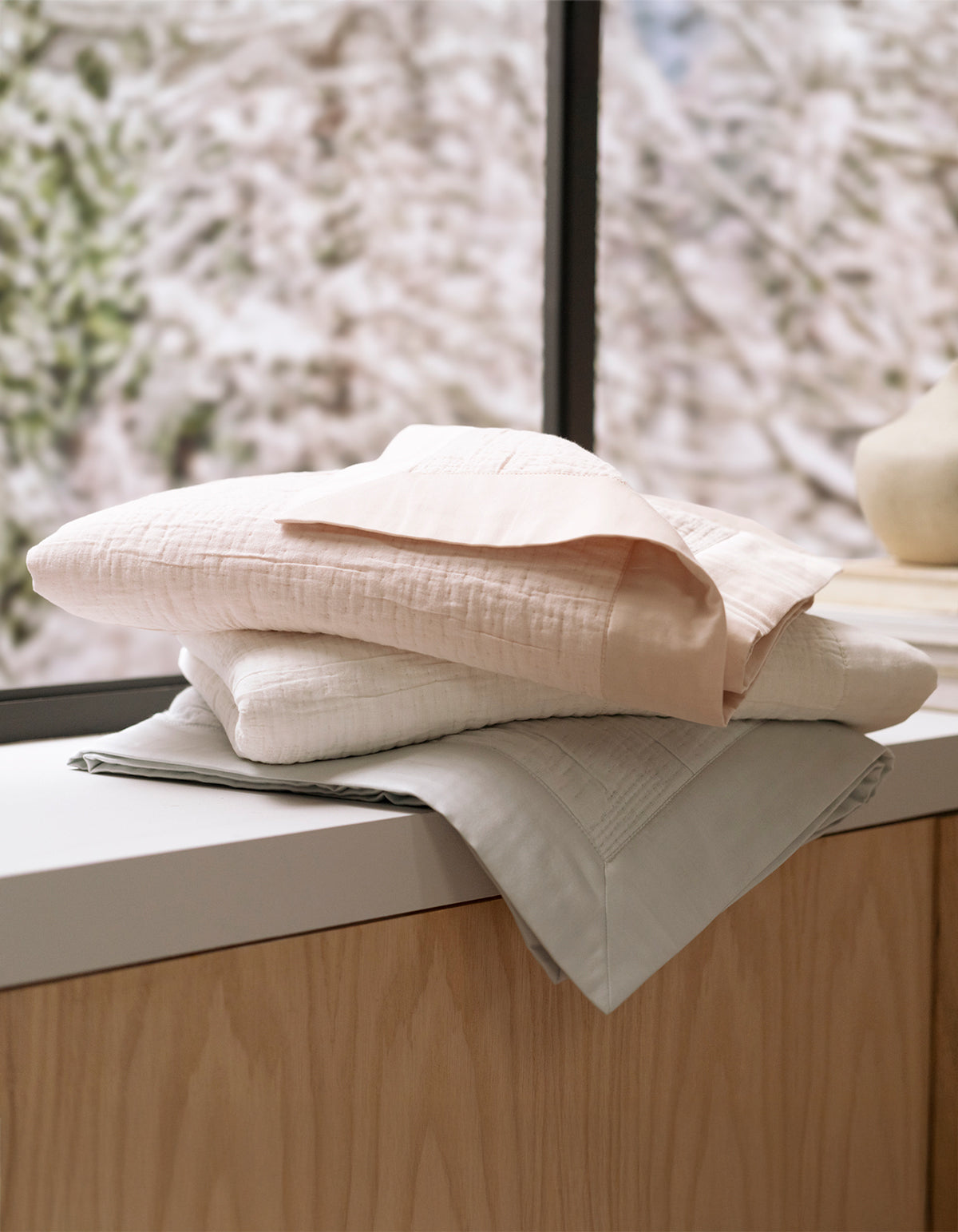 A stack of Cozy Earth's Little Bee Matelasse Baby Blankets in pastel colors rests on a wooden surface near a window, with a softly blurred snowy scene outside.|Color:Petal