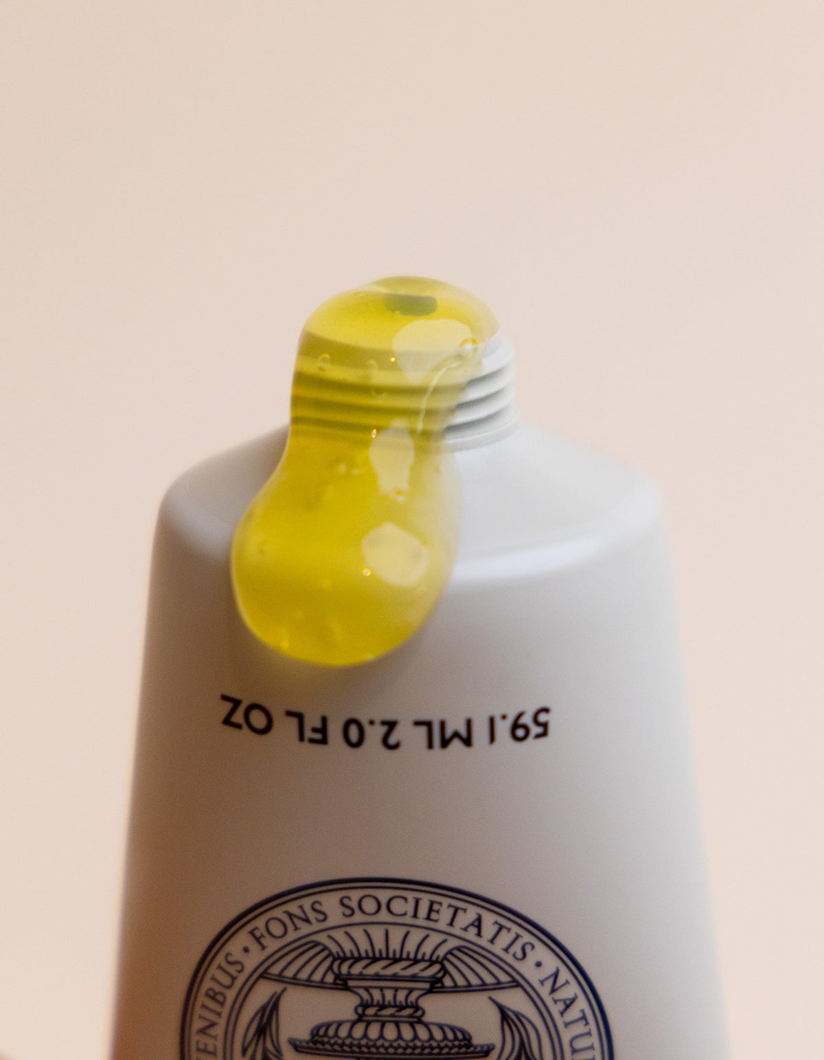 A close-up of the Cozy Earth Enzyme Mask: a white tube with black text and a circular emblem near the bottom, dispensing yellow gel. The softly lit background is plain.