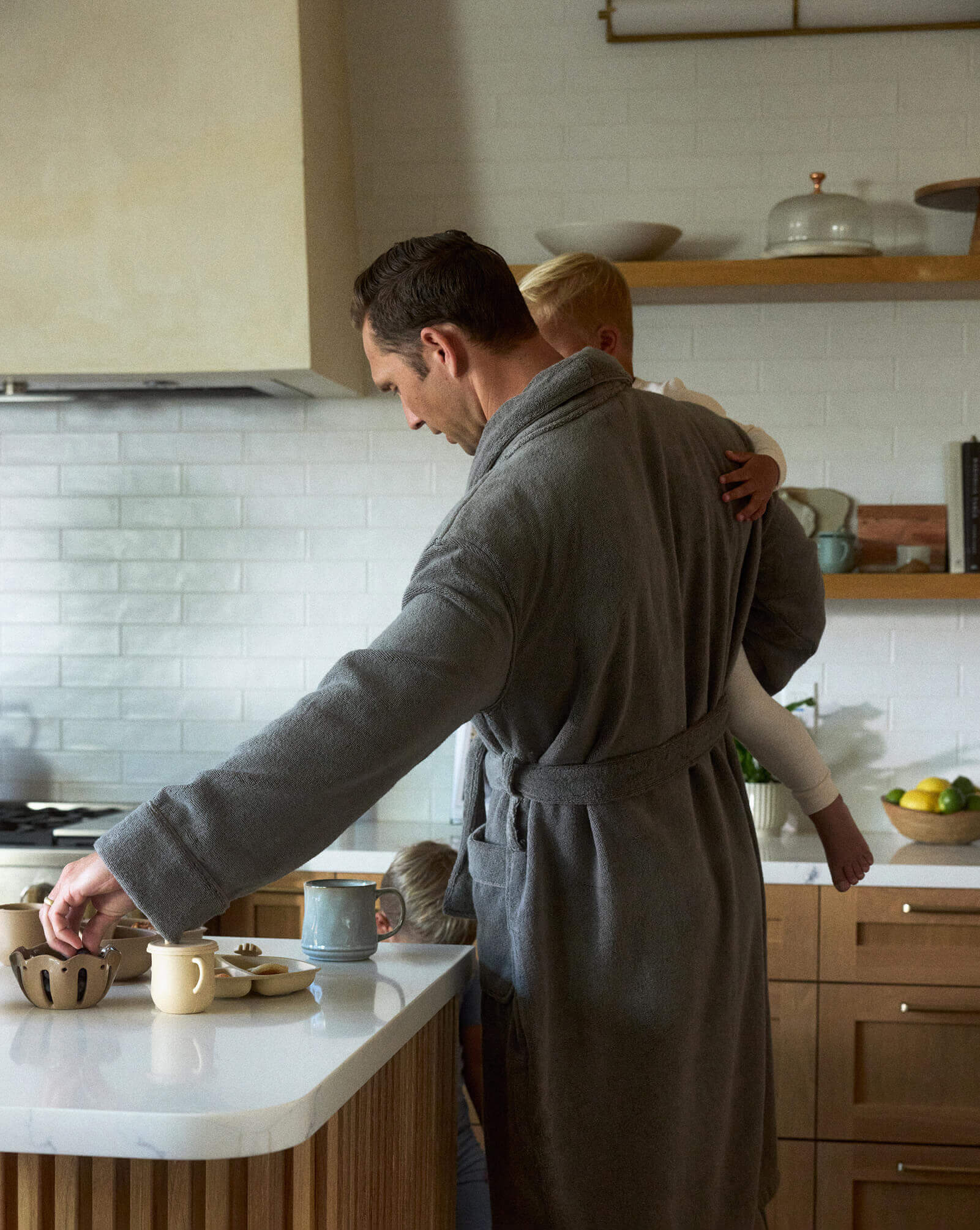 Man in a gray robe standing in a kitchen with a child on his shoulder.