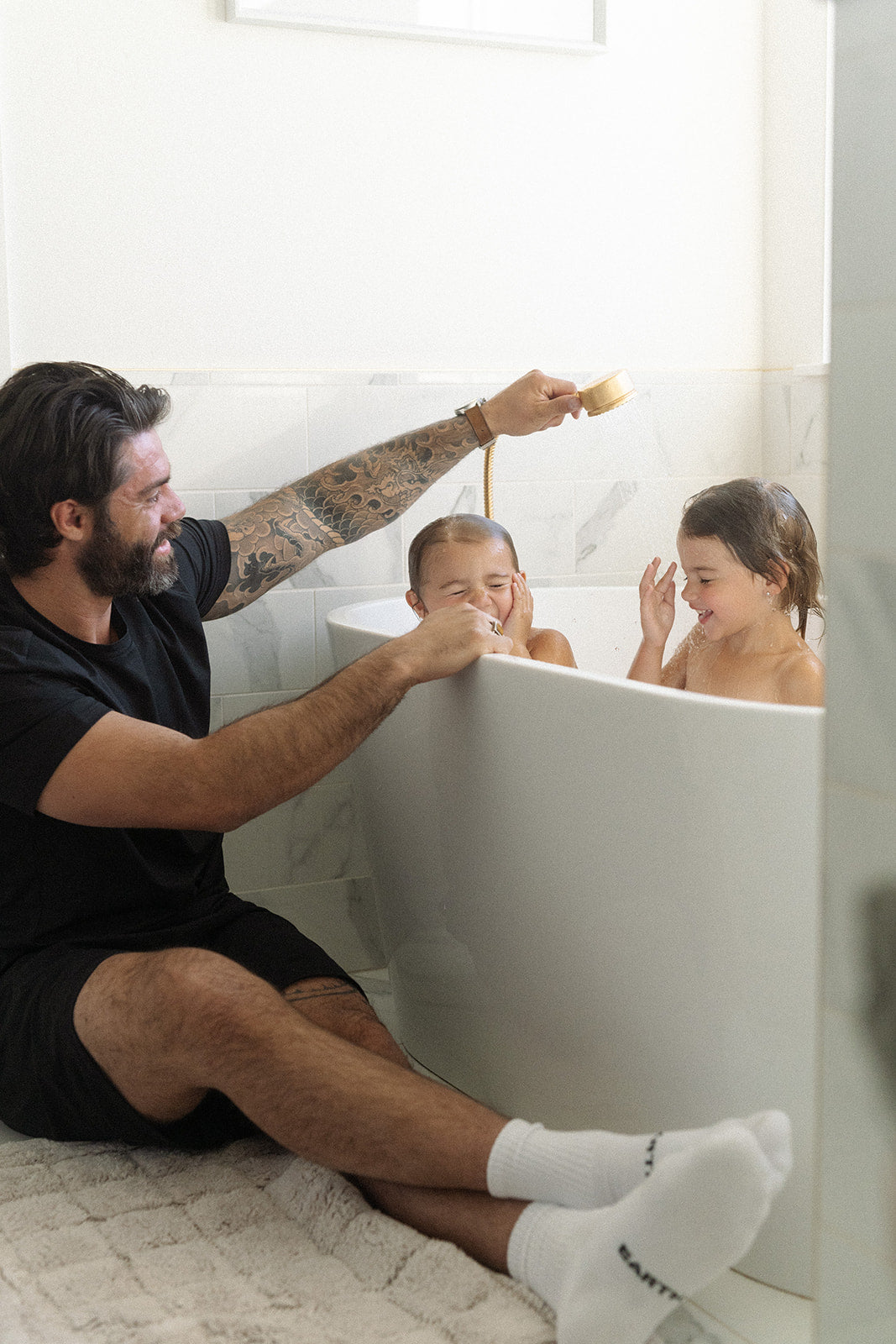 Man with tattoos and a beard sitting on the floor next to a bathtub with two children, one of whom is playing with a toy.
