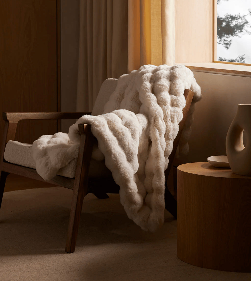 White textured blanket draped over a wooden chair in a cozy room with a window and side table.