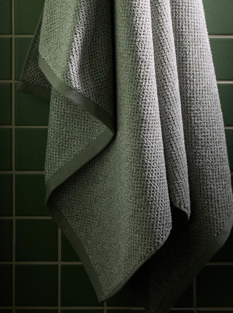 A close up shot of the Nantucket Towel hanging from a hook shows its soft and cozy texture. There is a tiled green wall behind it.