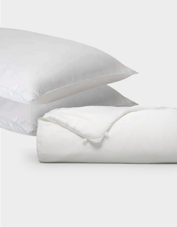Two white pillows from Cozy Earth's Silk Insert Bundle are stacked diagonally behind a rolled-up white duvet with a visible tie closure, all set against a plain light gray background.|Filling:Standard