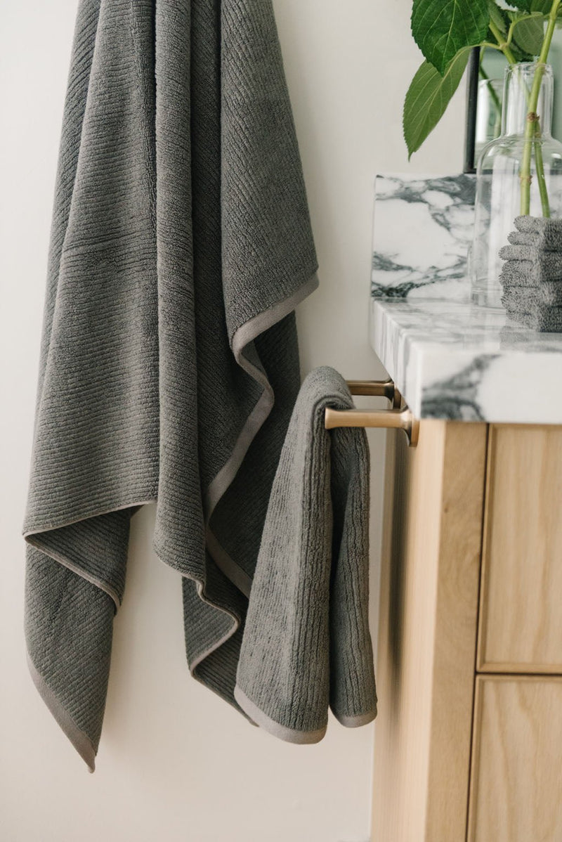 Ribbed Terry Hand Towels in the color charcoal. Photo of product taken with the product draped over a towel bar on the side of a white marble sink.