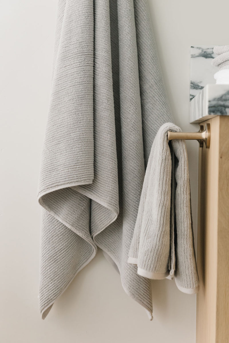Ribbed Terry Hand Towels in the color Light Grey. Photo of product taken with the product draped over a towel bar on the side of a white marble sink.
