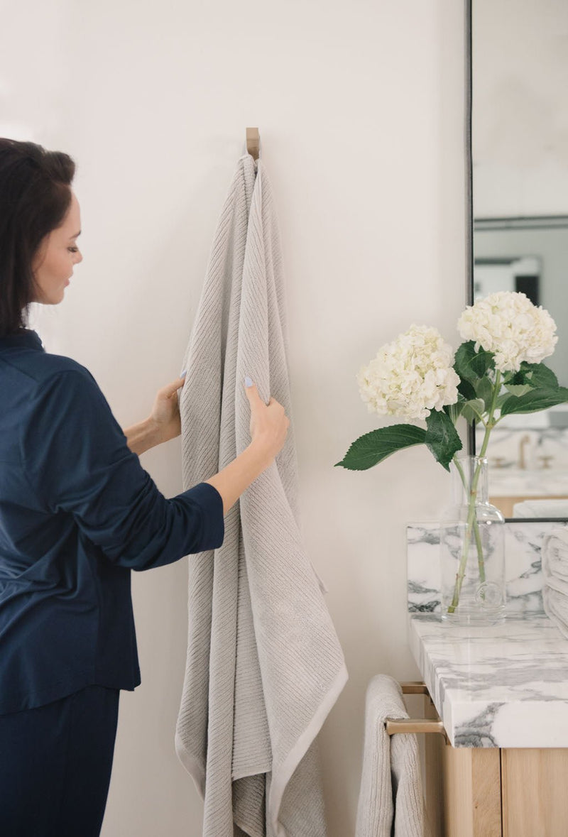 Ribbed Terry Bath Sheets in the color Light Grey. Photo of product taken in a bathroom next to a sink as a woman feels the towel.