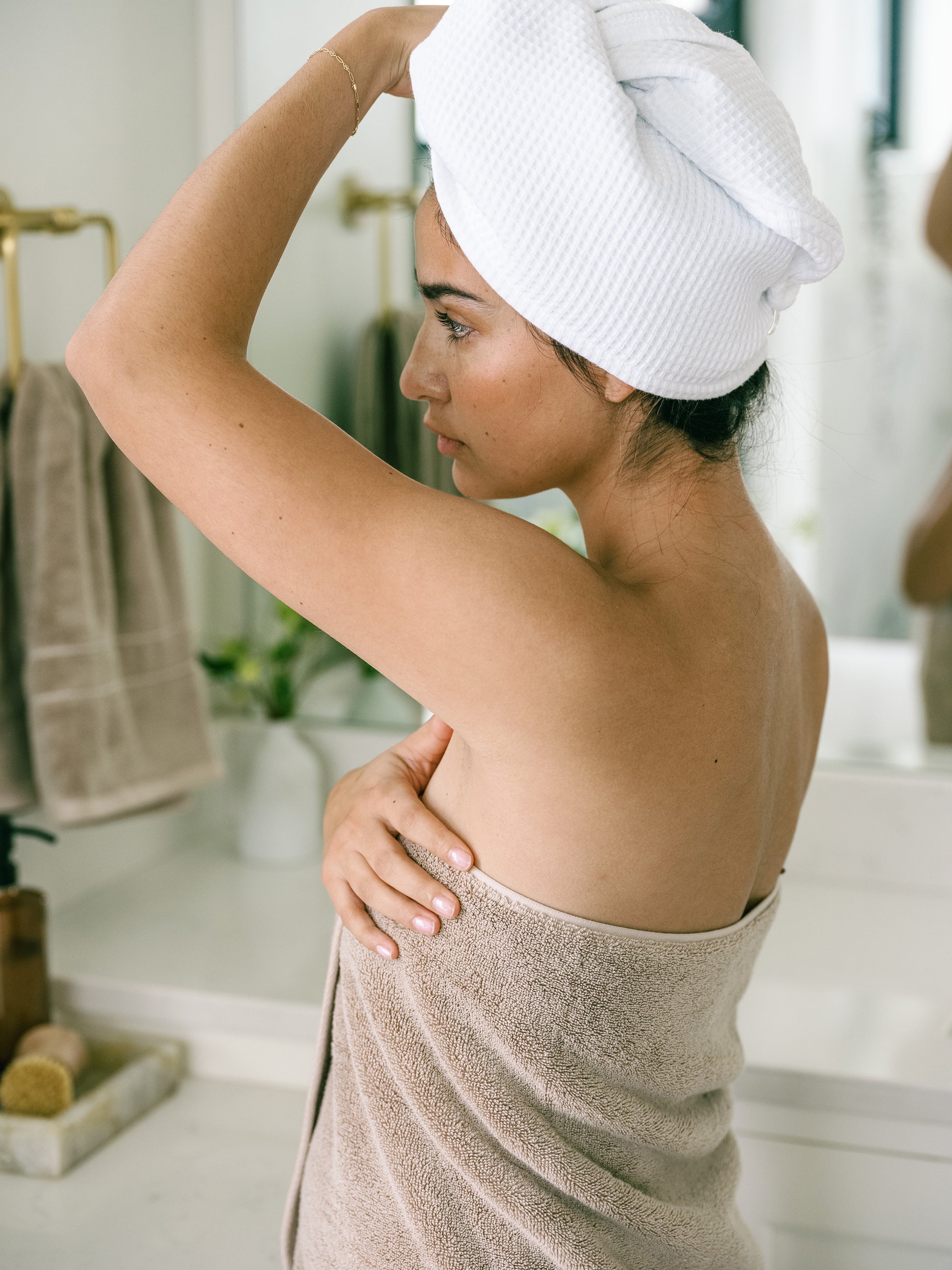 Premium Plush Bath Towel in the color Sand. Photo of Sand Premium Plush Bath Towel taken in a bathroom with a white tile backsplash. There is a brunette woman holding the towel close to her body as she looks in the mirror. 