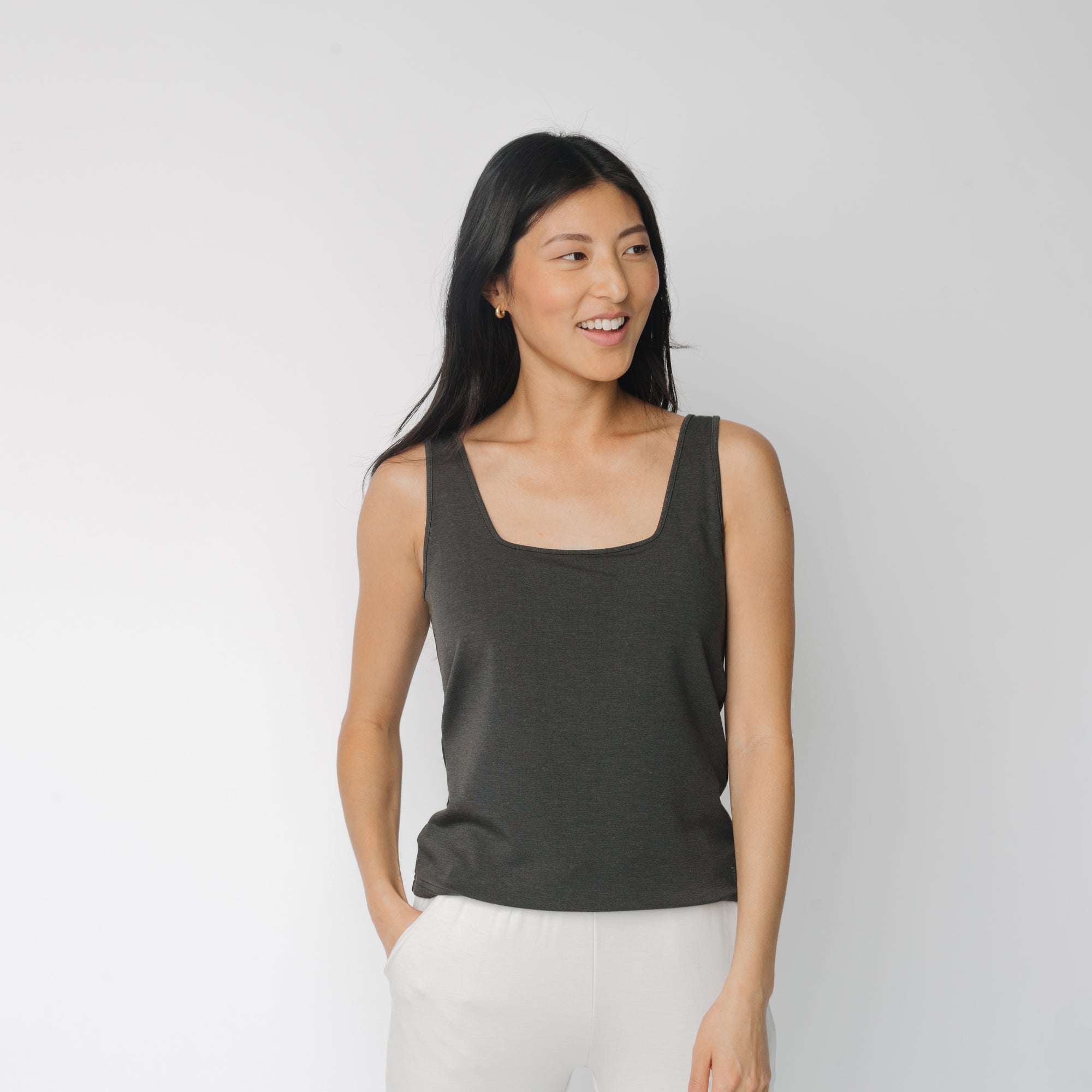 A person with long dark hair smiles while looking to the side. They are wearing a Cozy Earth Women's Ultra-Soft Bamboo Square Neck Tank in dark gray and light-colored pants, standing against a plain white background with one hand in their pocket. 