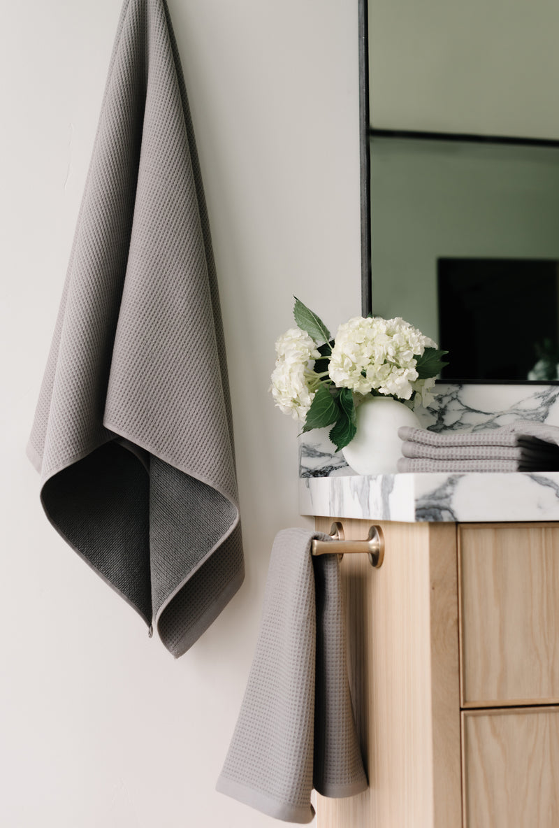 Charcoal Waffle Hand Towel hanging from a gold towel bar. The photo was taken at a white marble bathroom sink. A Waffle Bath Towel is seen hanging on the wall in the background.