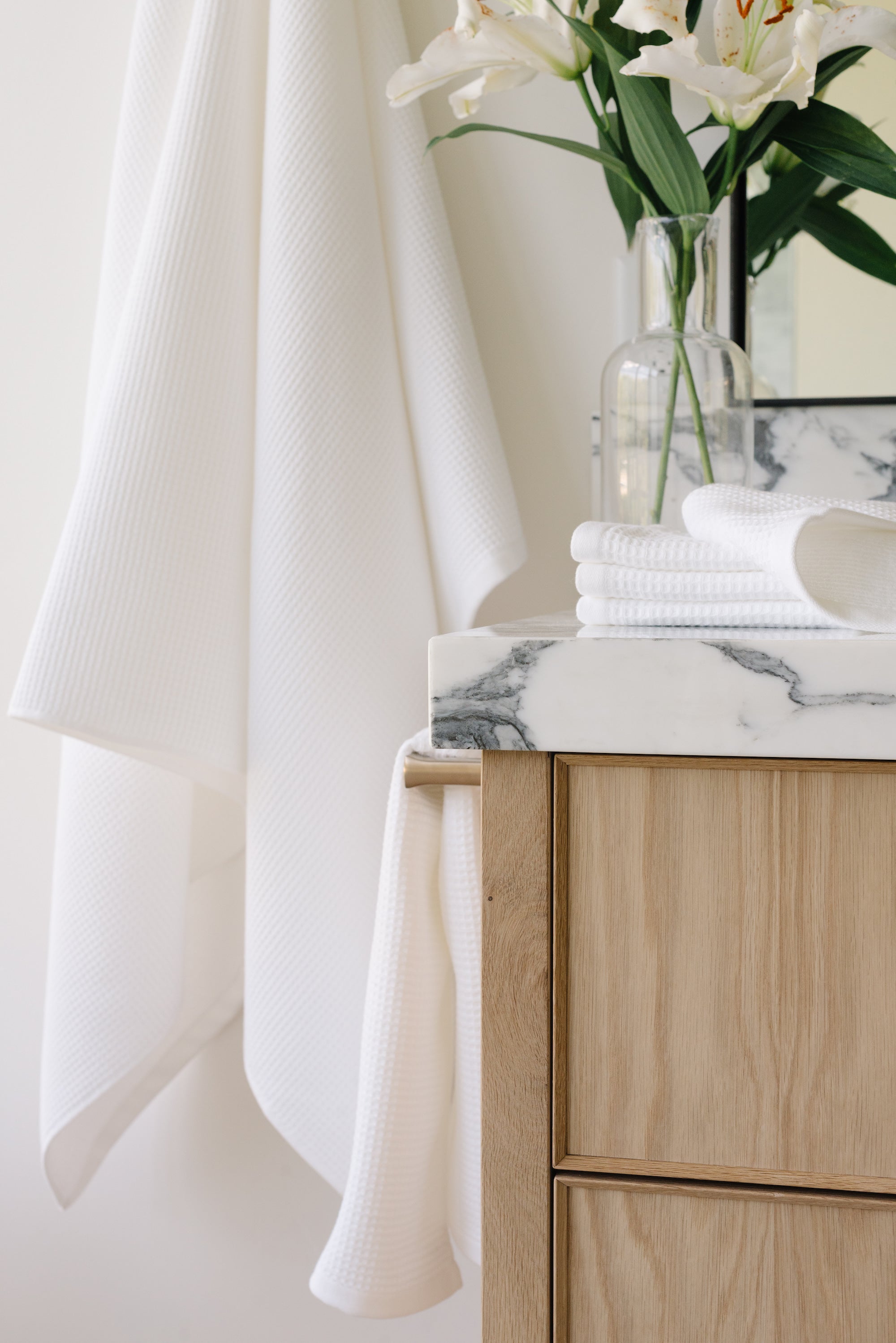 White Waffle Hand Towel hanging from a gold towel bar. The photo was taken at a white marble bathroom sink. A Waffle Bath Towel is seen hanging on the wall in the background. 