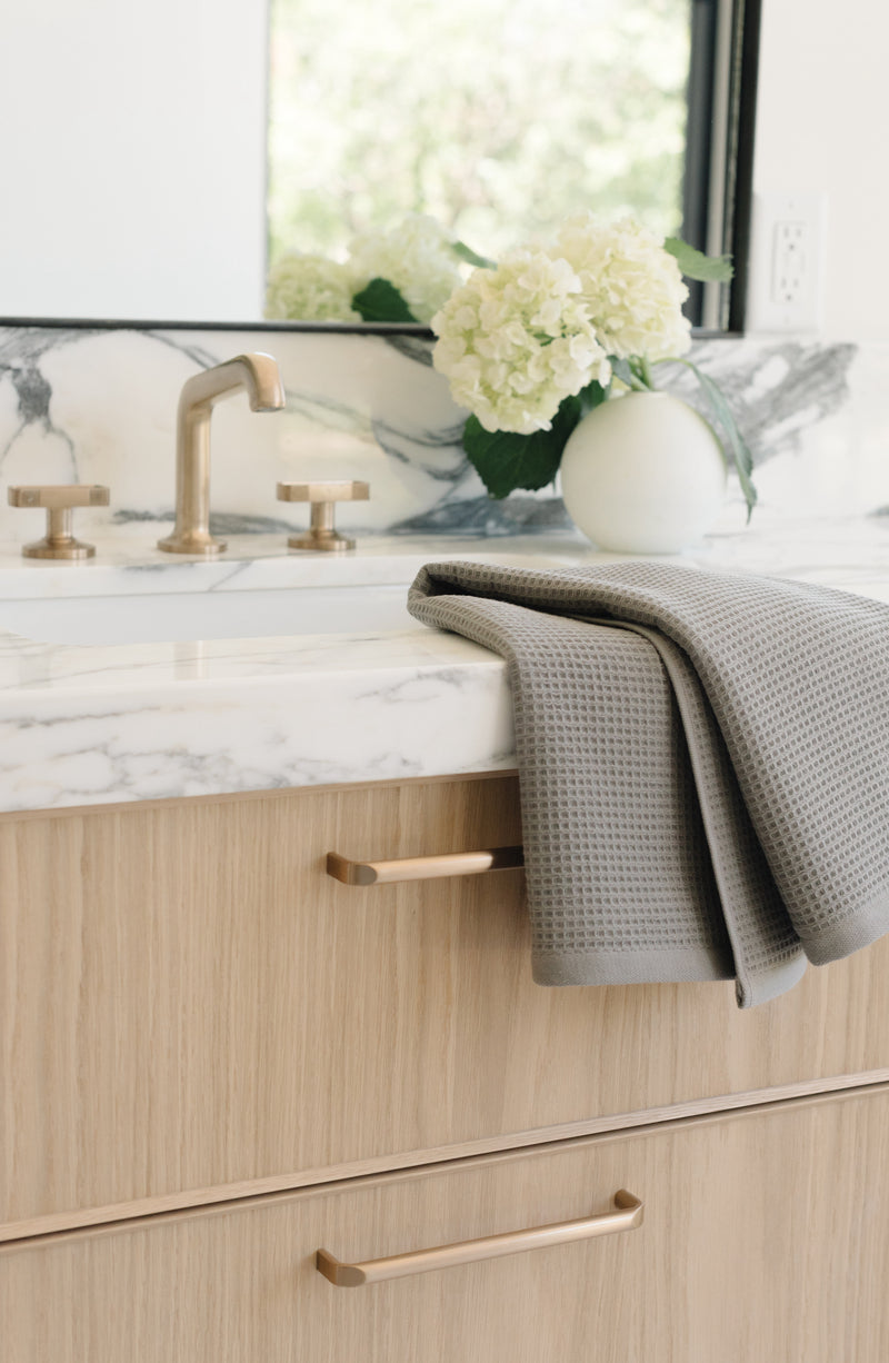 Charcoal Waffle Hand Towel resting folded on a white marble bathroom sink. The towel is folded so that different views of the towel can be seen.